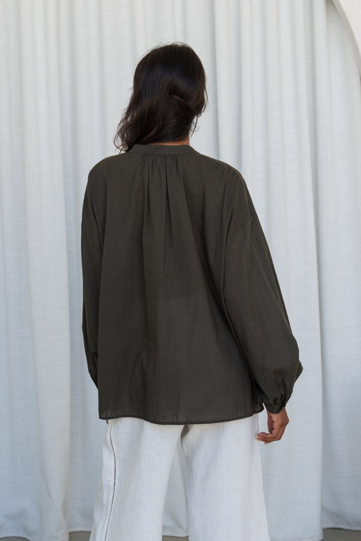 LILLY PILLY woman wearing a khaki Ember cotton voile shirt and ivory linen pants against a curtain background showing back view
