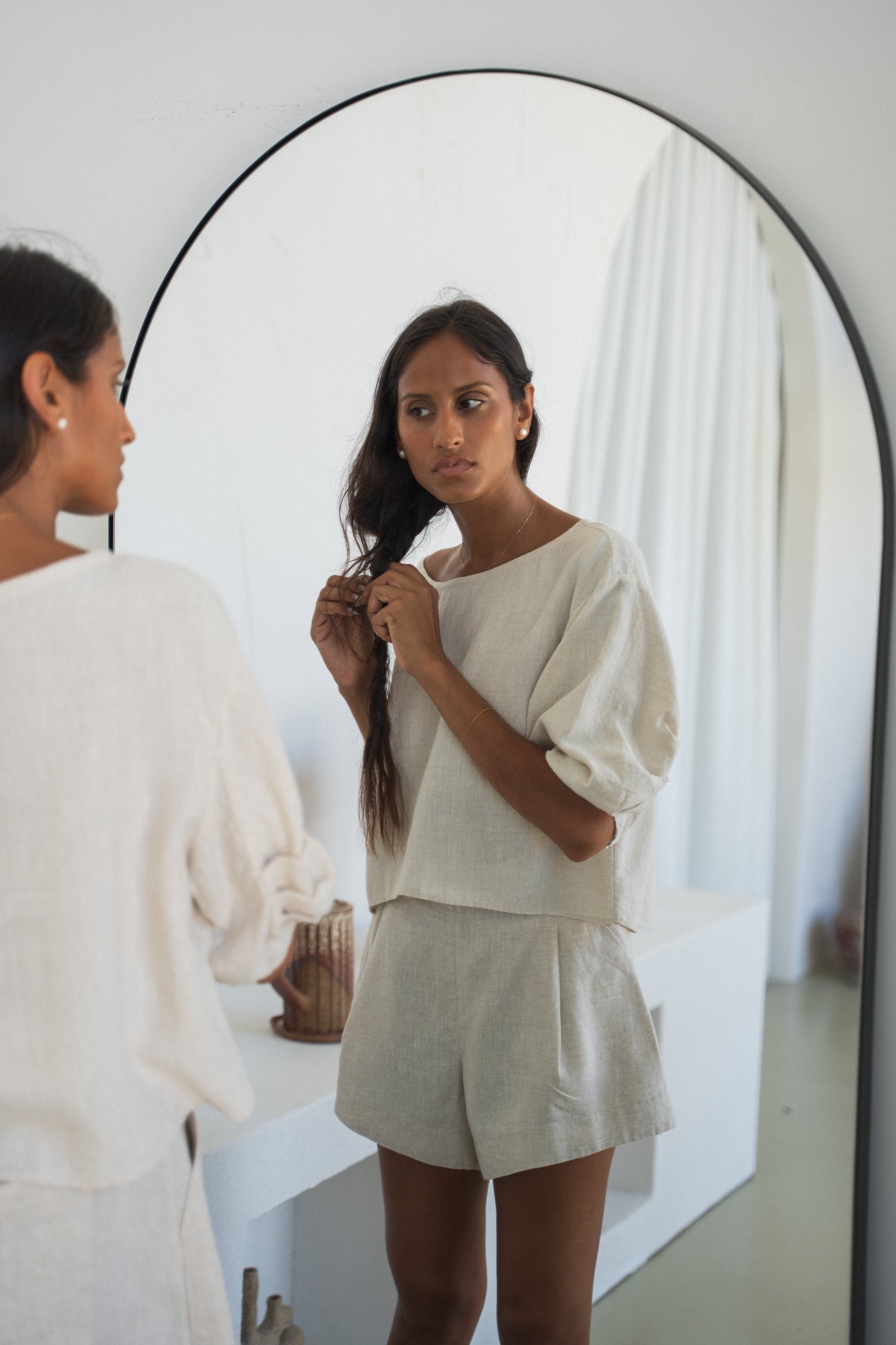Woman adjusting her hair in front of a mirror wearing oatmeal Leia linen top against a light gray background