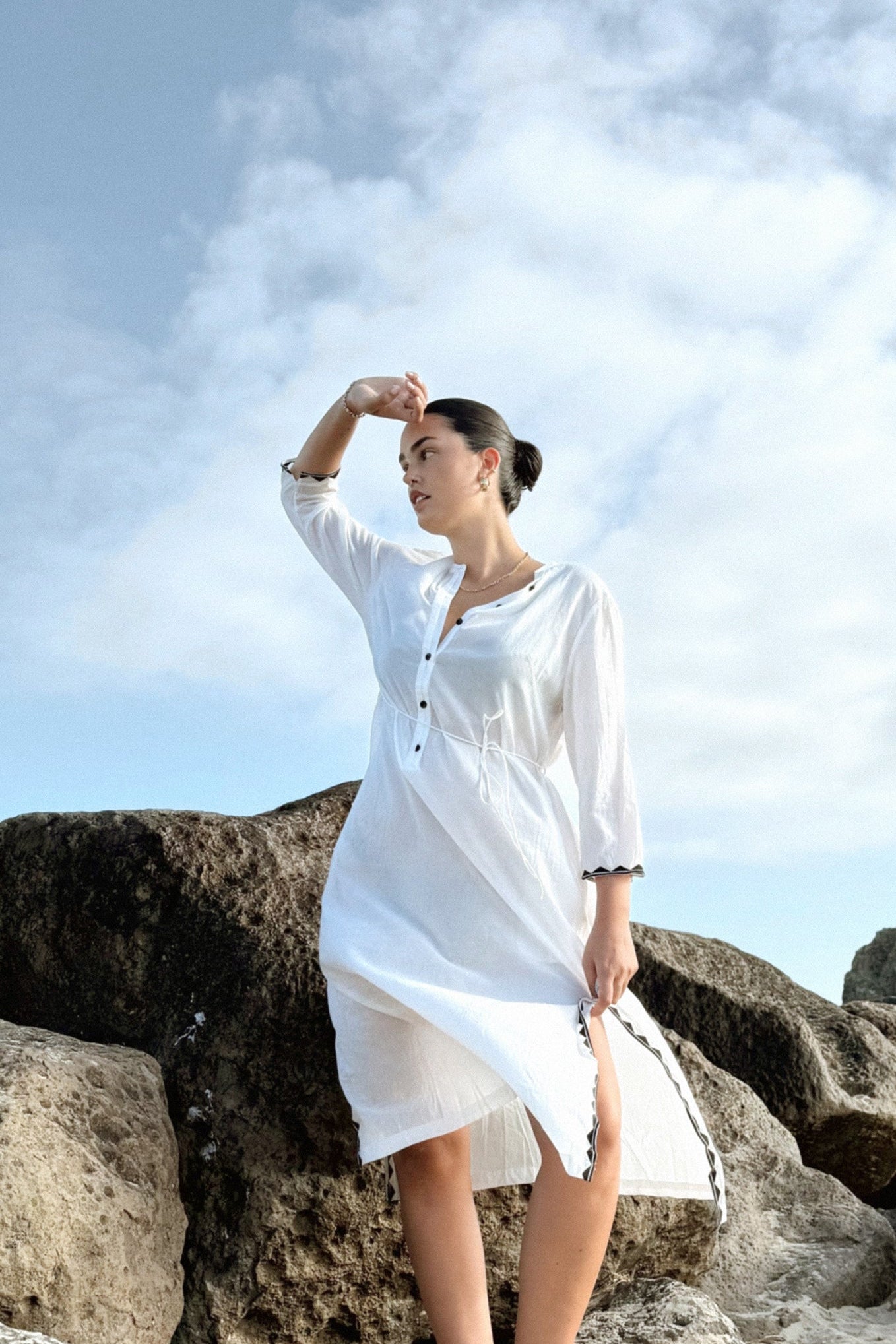 LILLY PILLY woman in a ivory Callie cotton dress standing on rocks with a blue sky background