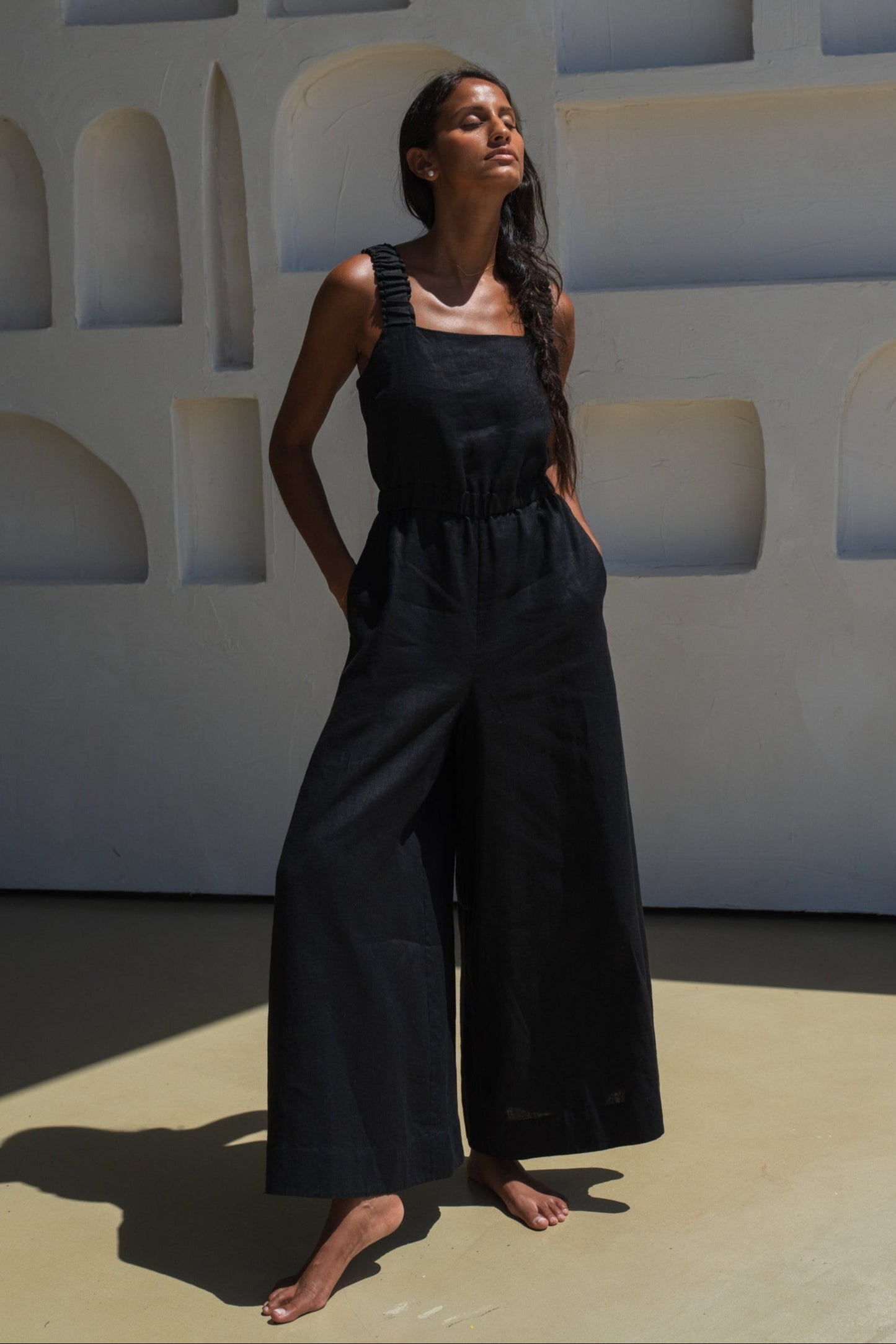 LILLY PILLY woman wearing a black Keely linen jumpsuit standing against a geometric wall.