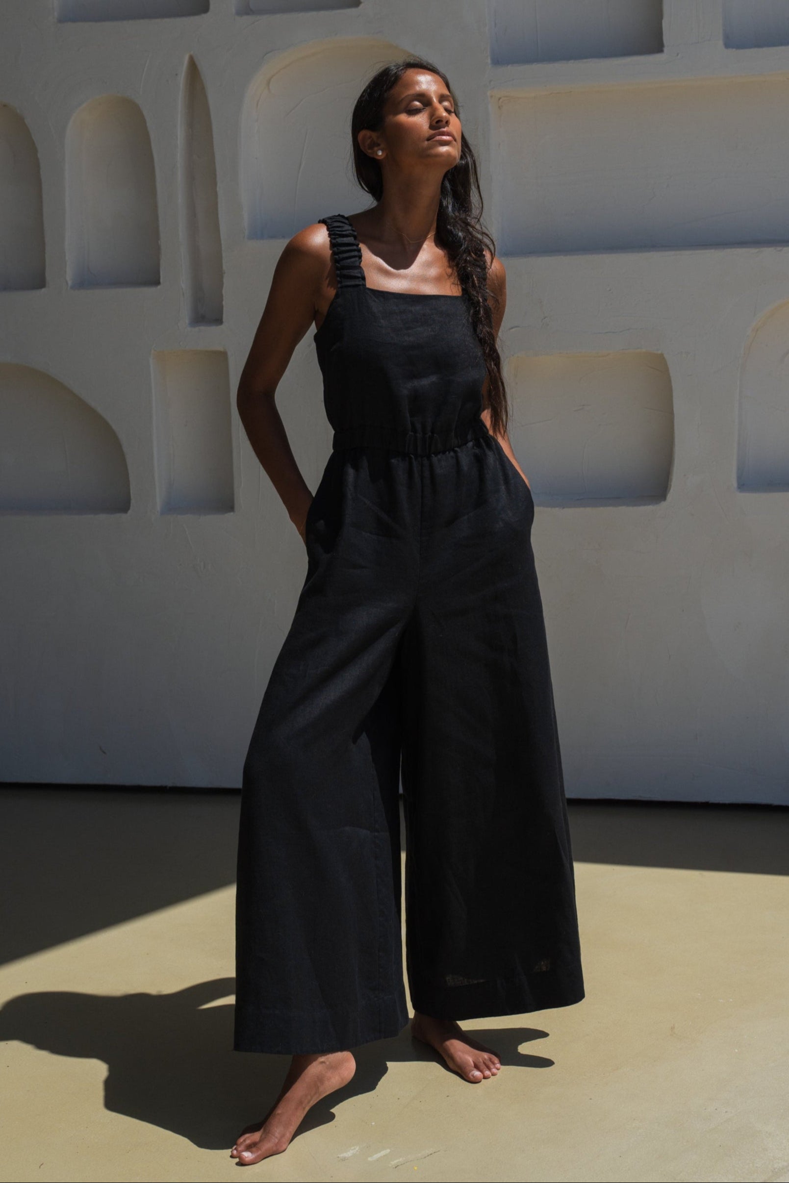 LILLY PILLY woman wearing a black Keely linen jumpsuit standing against a geometric wall.