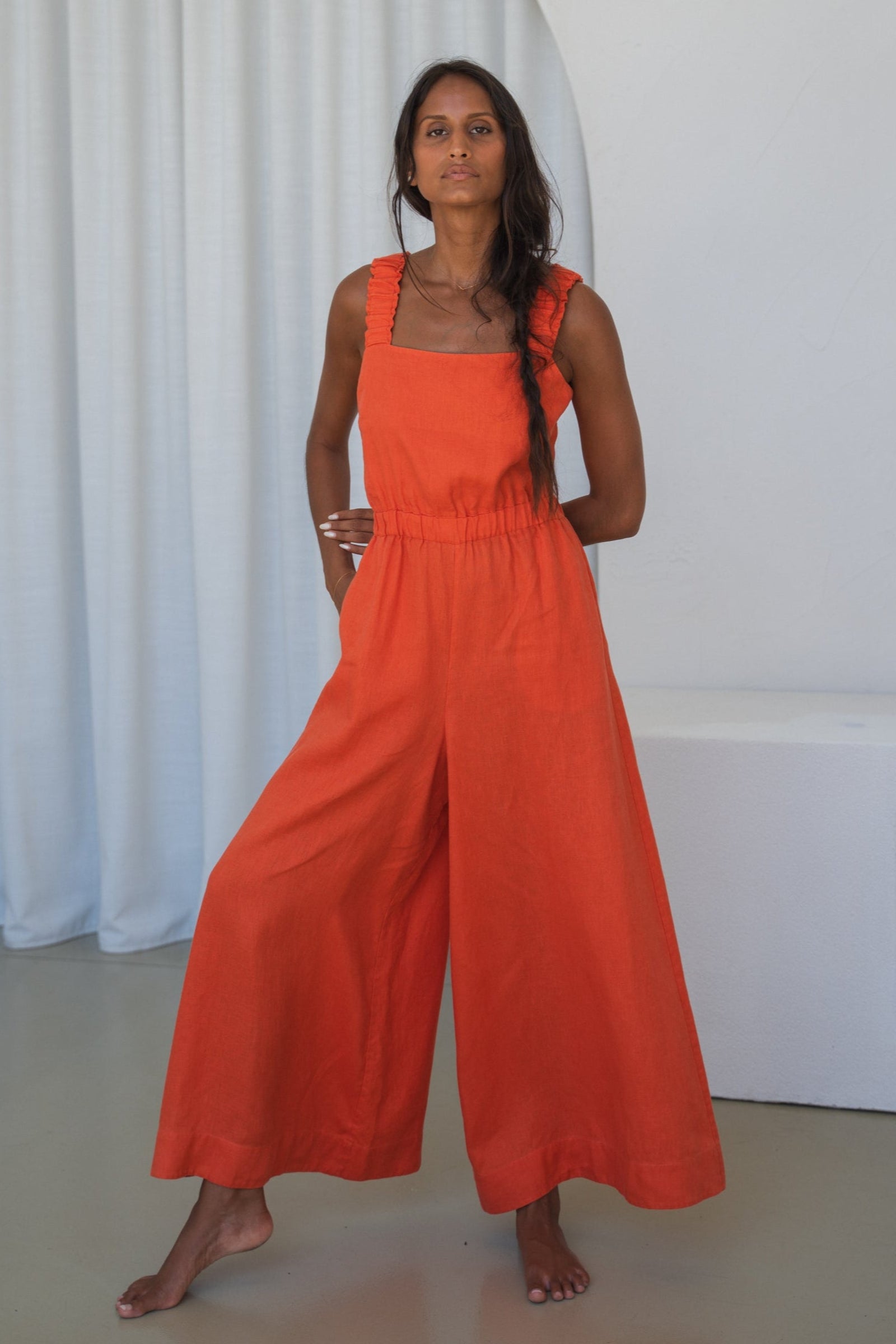 LILLY PILLY woman in an spritz Keely linen jumpsuit standing against a white curtain background