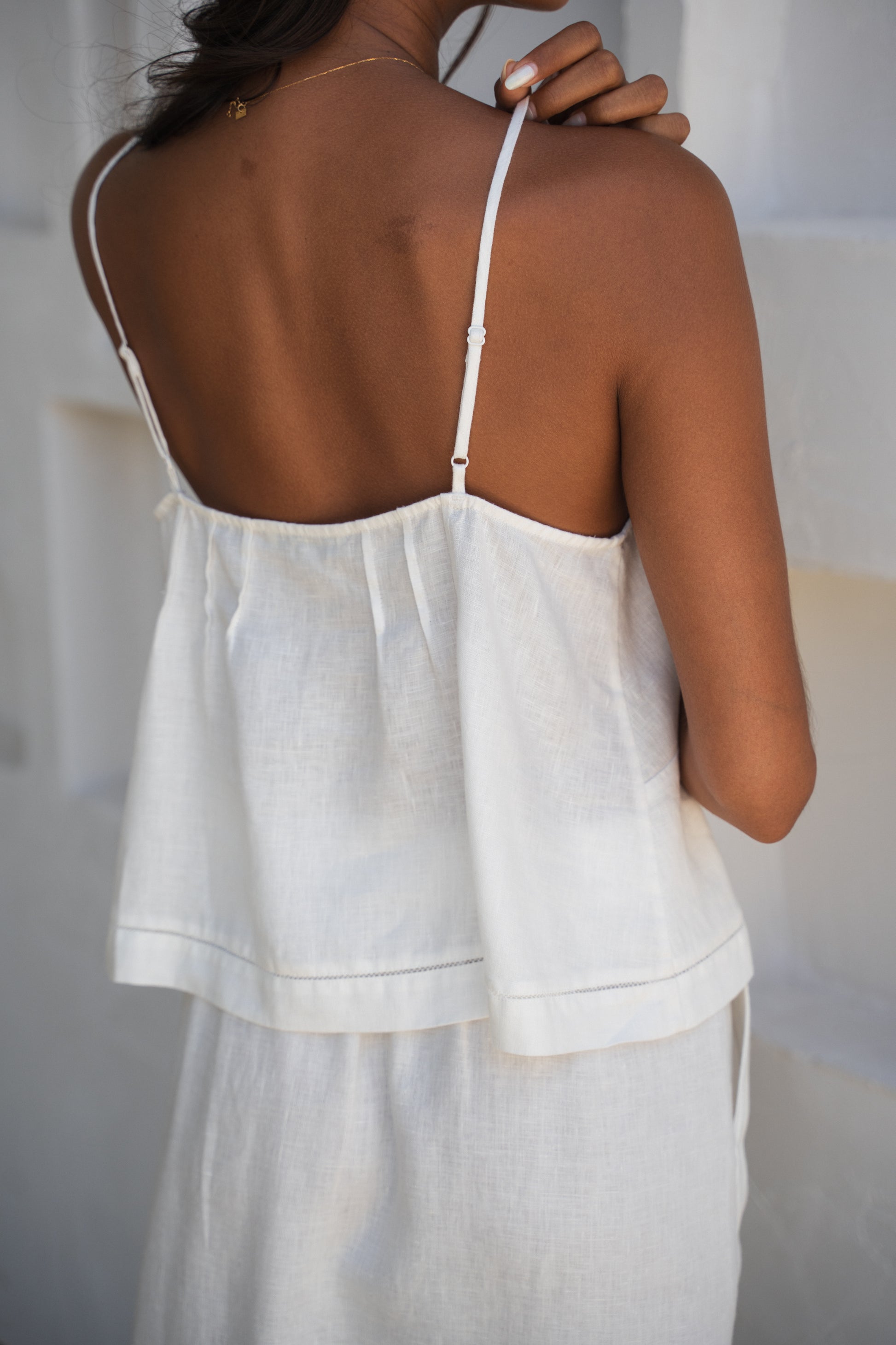 Close up back view of LILLY PILLY woman wearing a ivory Bea linen cami top standing against a textured white wall.