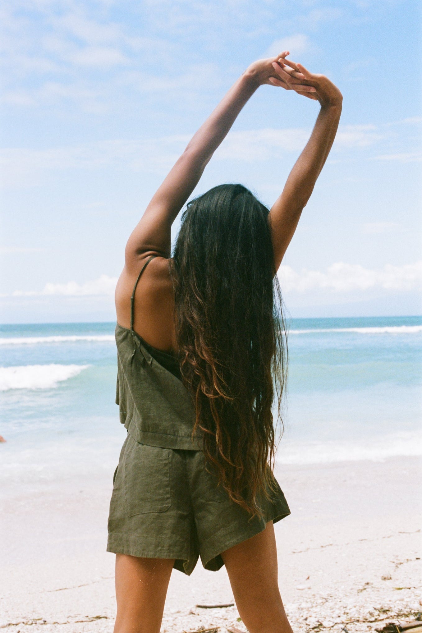 LILLY PILLY woman wearing a khaki Bea linen cami top and Gaia shorts outfit on a beach showing the back view while stretching on a beach with ocean view