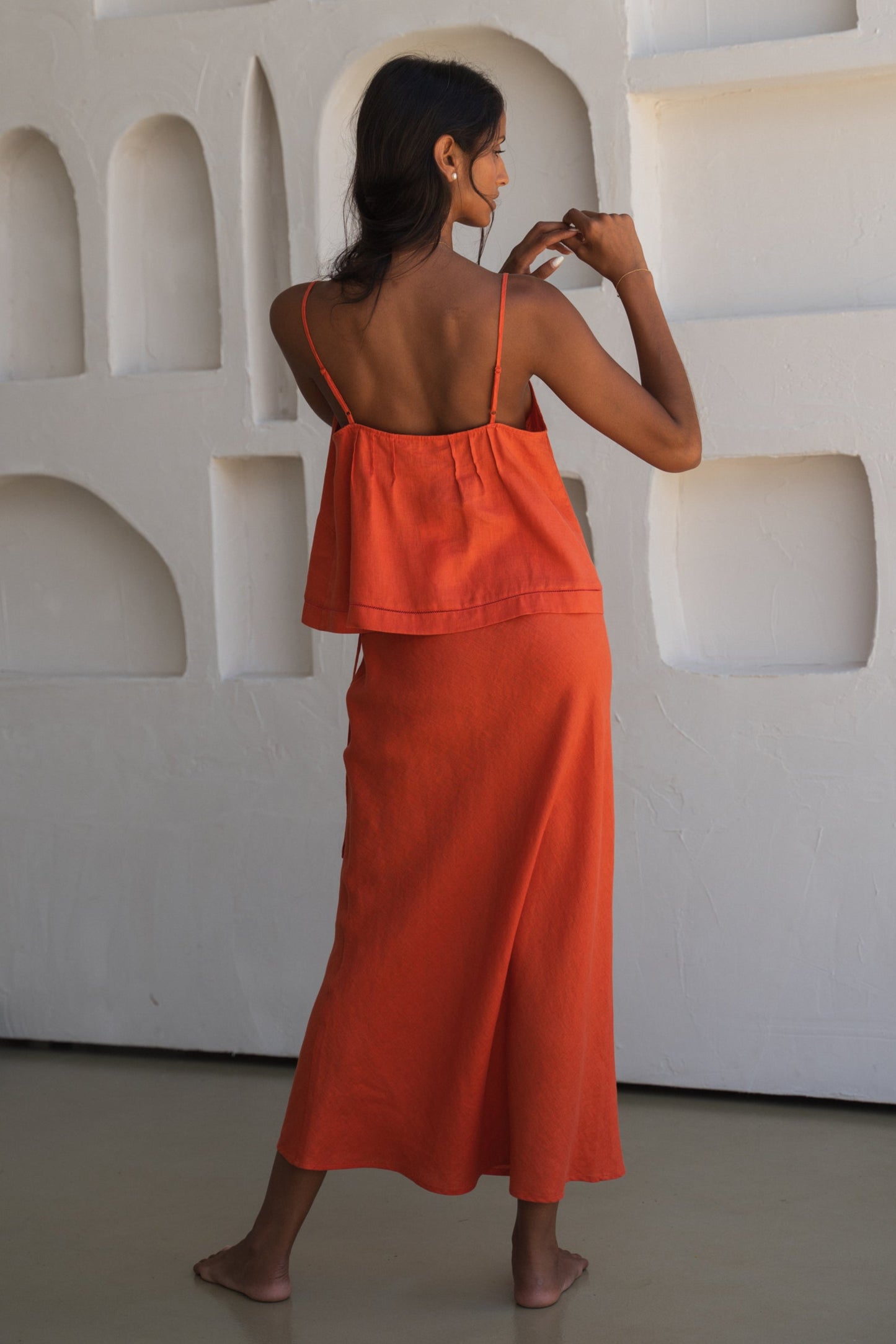 Close up of LILLY PILLY woman wearing a spritz Bea linen cami top with thin straps showing back view while posing against a textured white wall.