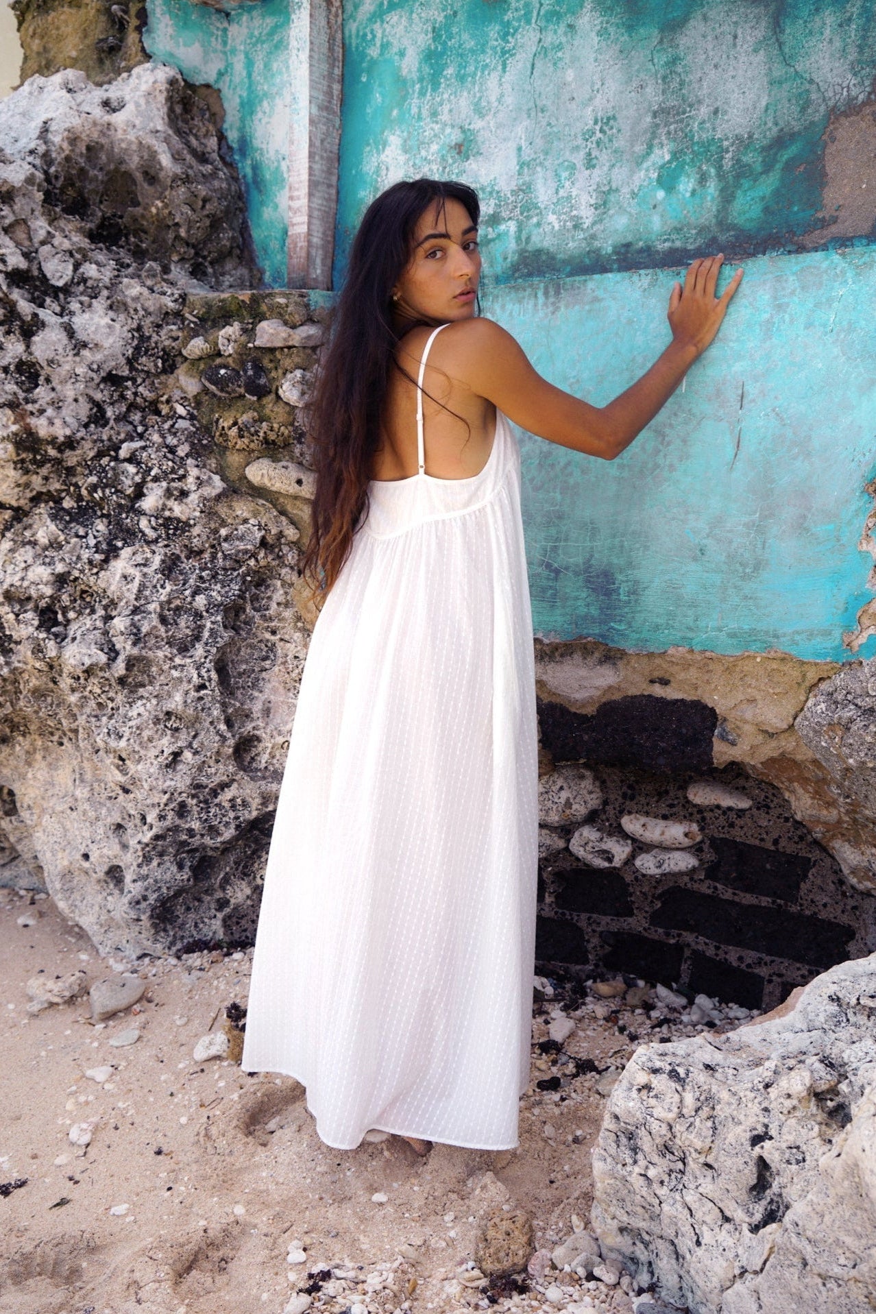 Woman in a white dress standing against a textured wall with peeling paint.
