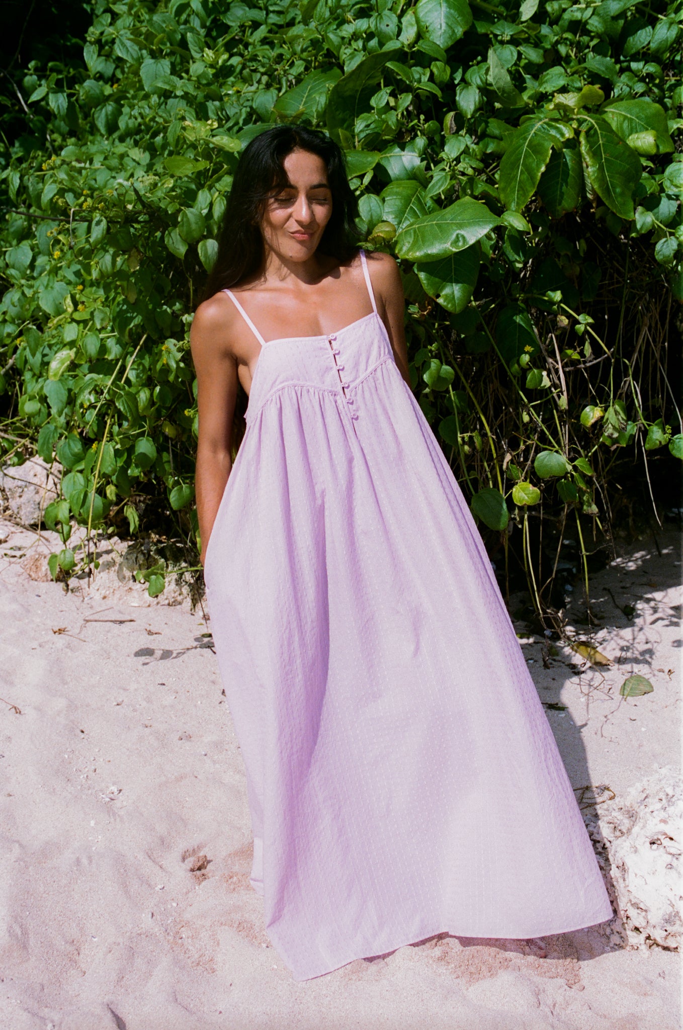 Woman in a long light purple dress standing in a natural setting with greenery.