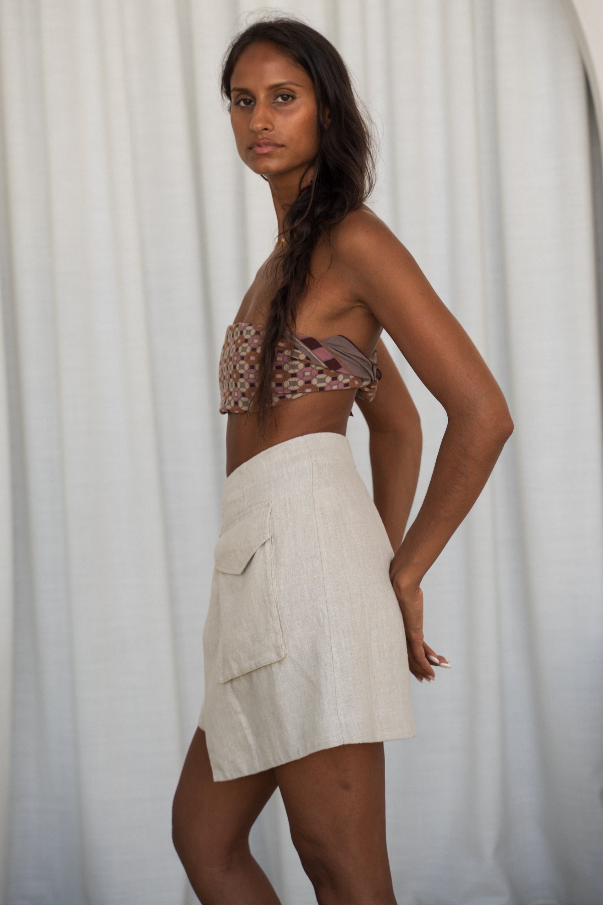Woman wearing a patterned top and oatmeal Betty linen skirt against a white curtain background