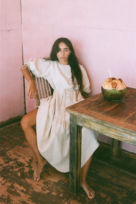 LILLY PILLY woman in an oatmeal Charlotte dress sitting on a chair in a room with pink walls and a wooden table.