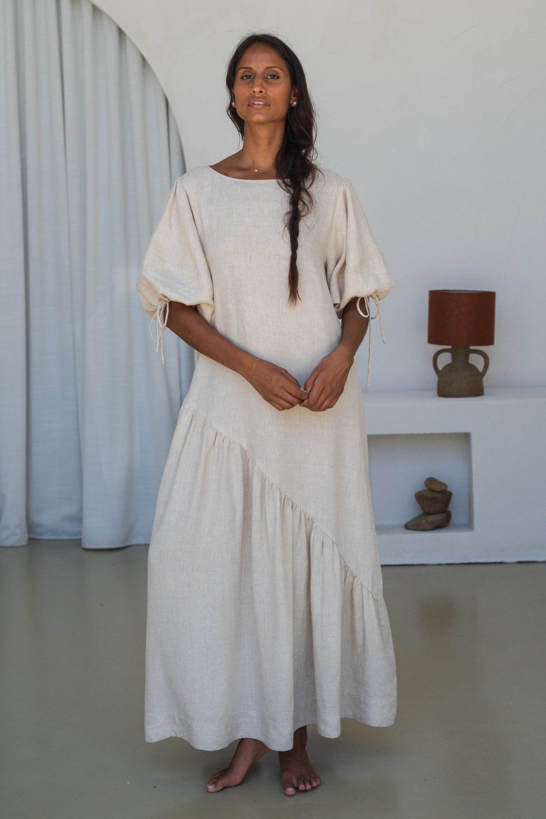 LILLY PILLY woman in an oatmeal Charlotte linen dress standing in a minimalistic room with a lamp and shelves.