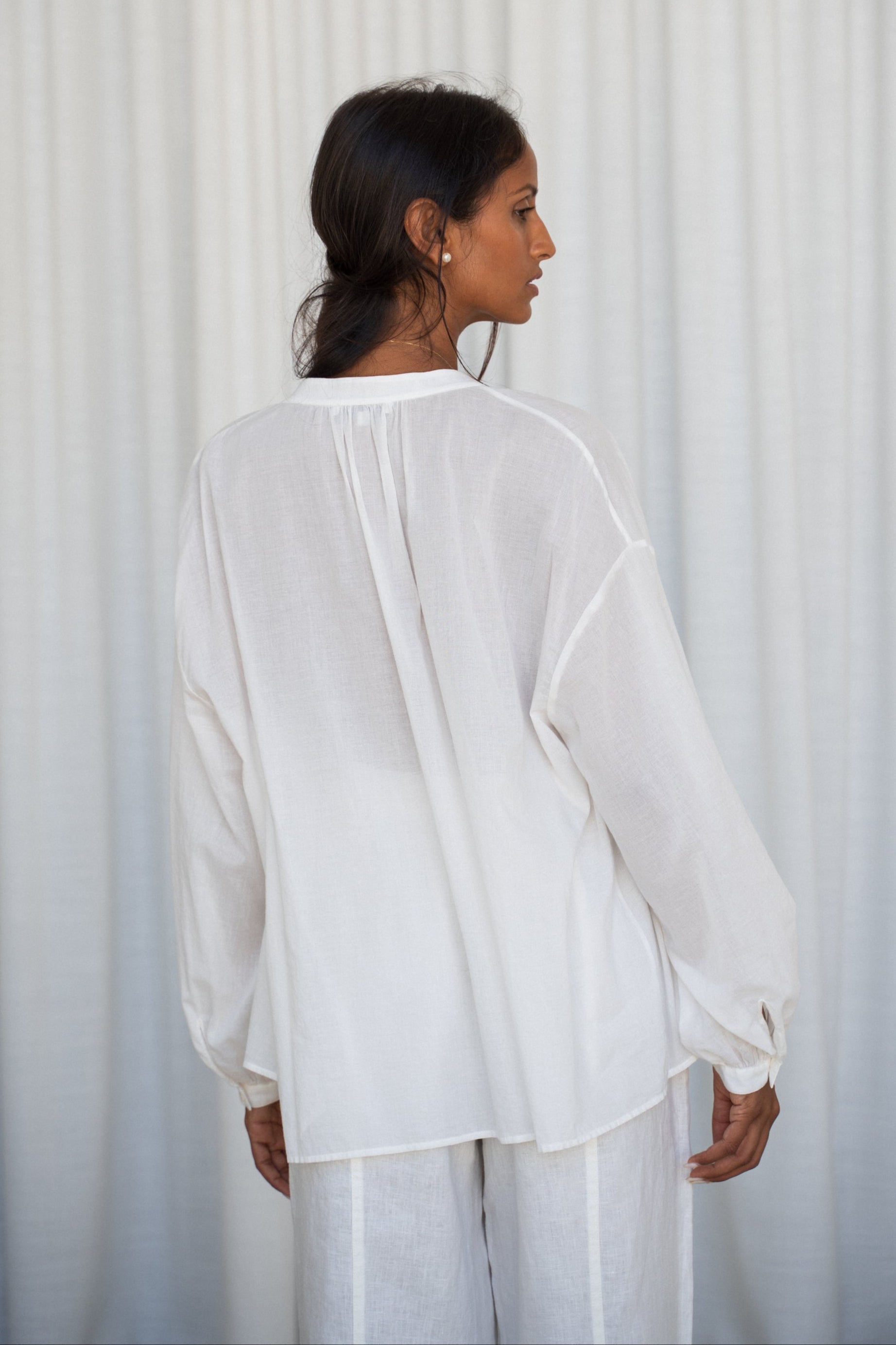 LILLY PILLY woman wearing a ivory Ember cotton voile shirt and ivory linen pants against a light curtain background showing back view