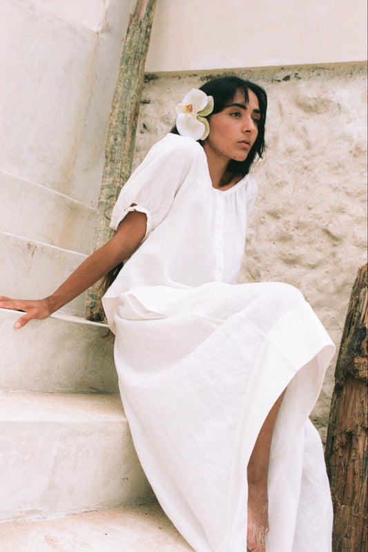LILLY PILLY woman in an ivory Hannah linen skirt and Pia linen top sitting on stone steps with a white flower in her hair.