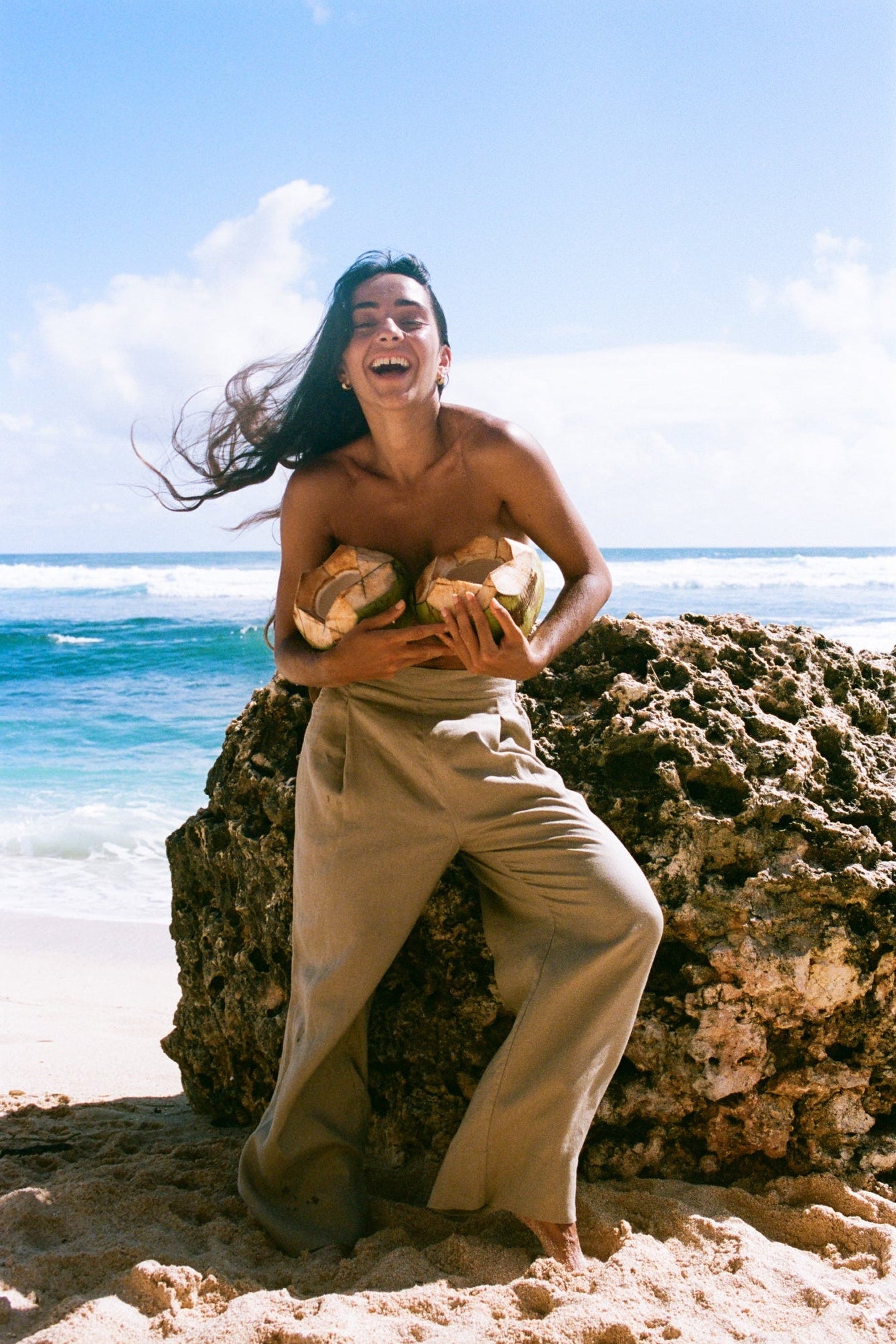 Person sitting on a rock by the ocean holding coconuts, wearing Ivy taupe linen pants with a clear blue sky.