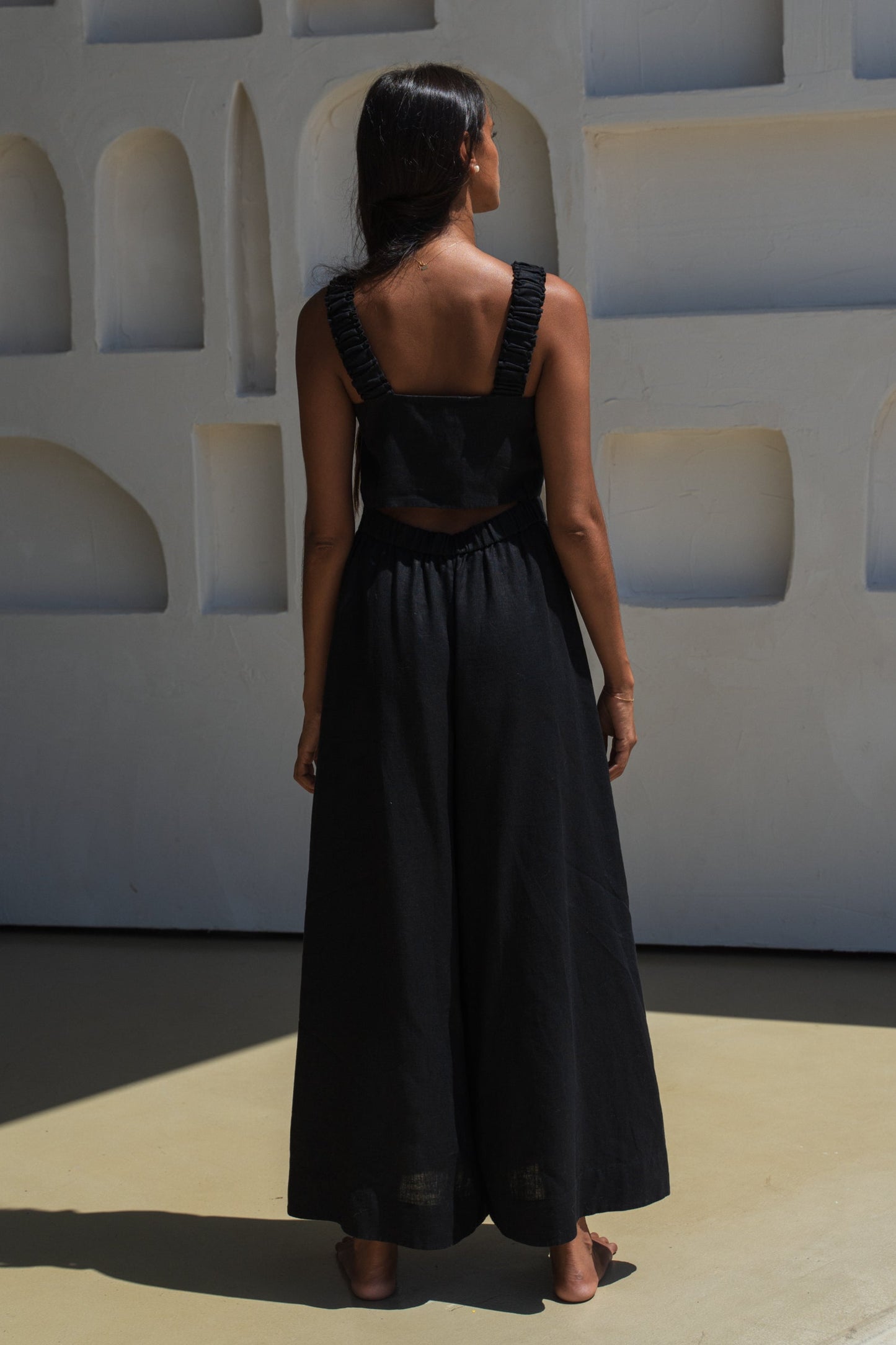 LILLY PILLY woman wearing a black Keely linen jumpsuit showing back view while standing against a geometric wall.