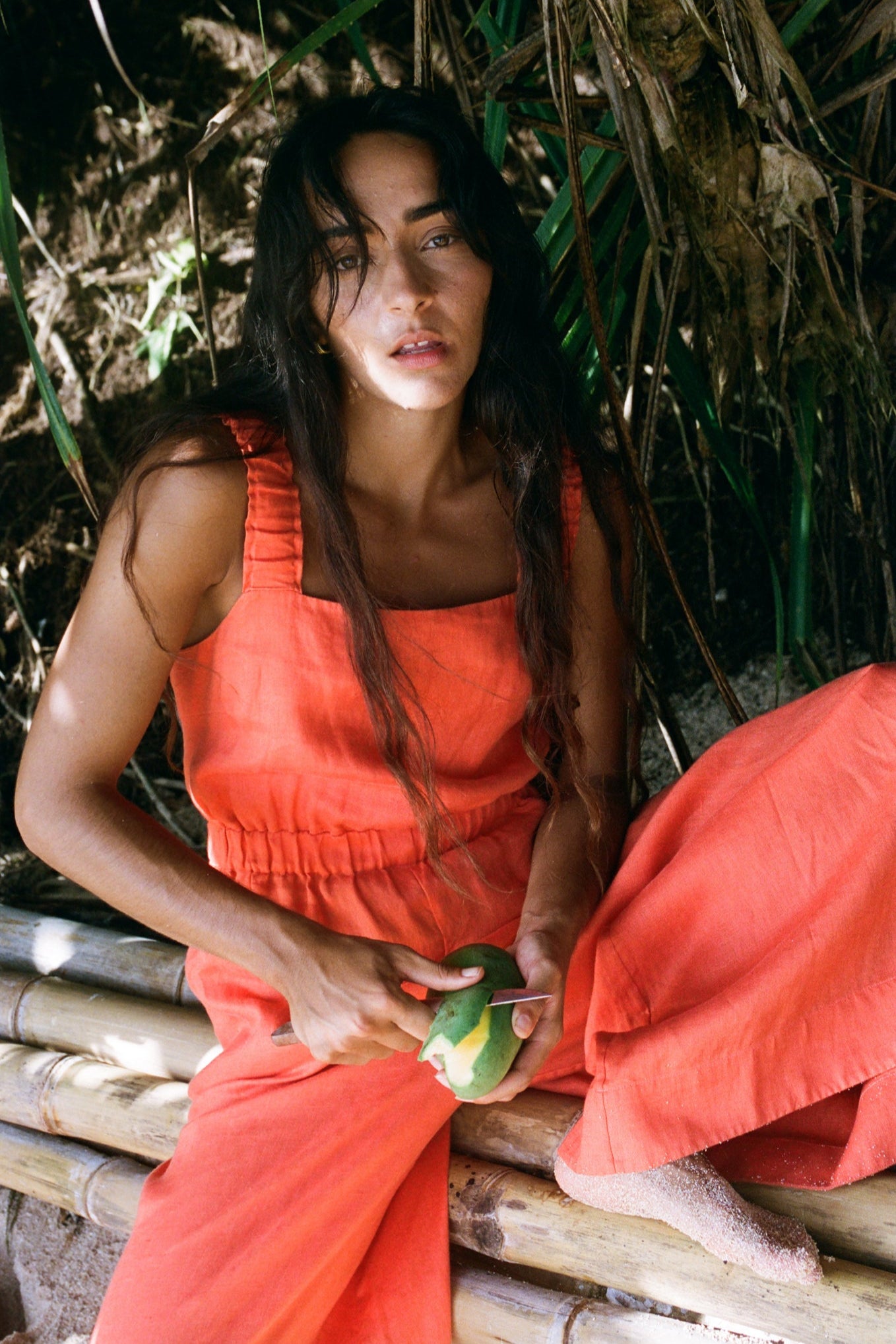 LILLY PILLY woman in an spritz Keely jumpsuit sitting on a bamboo structure with green leaves in the background