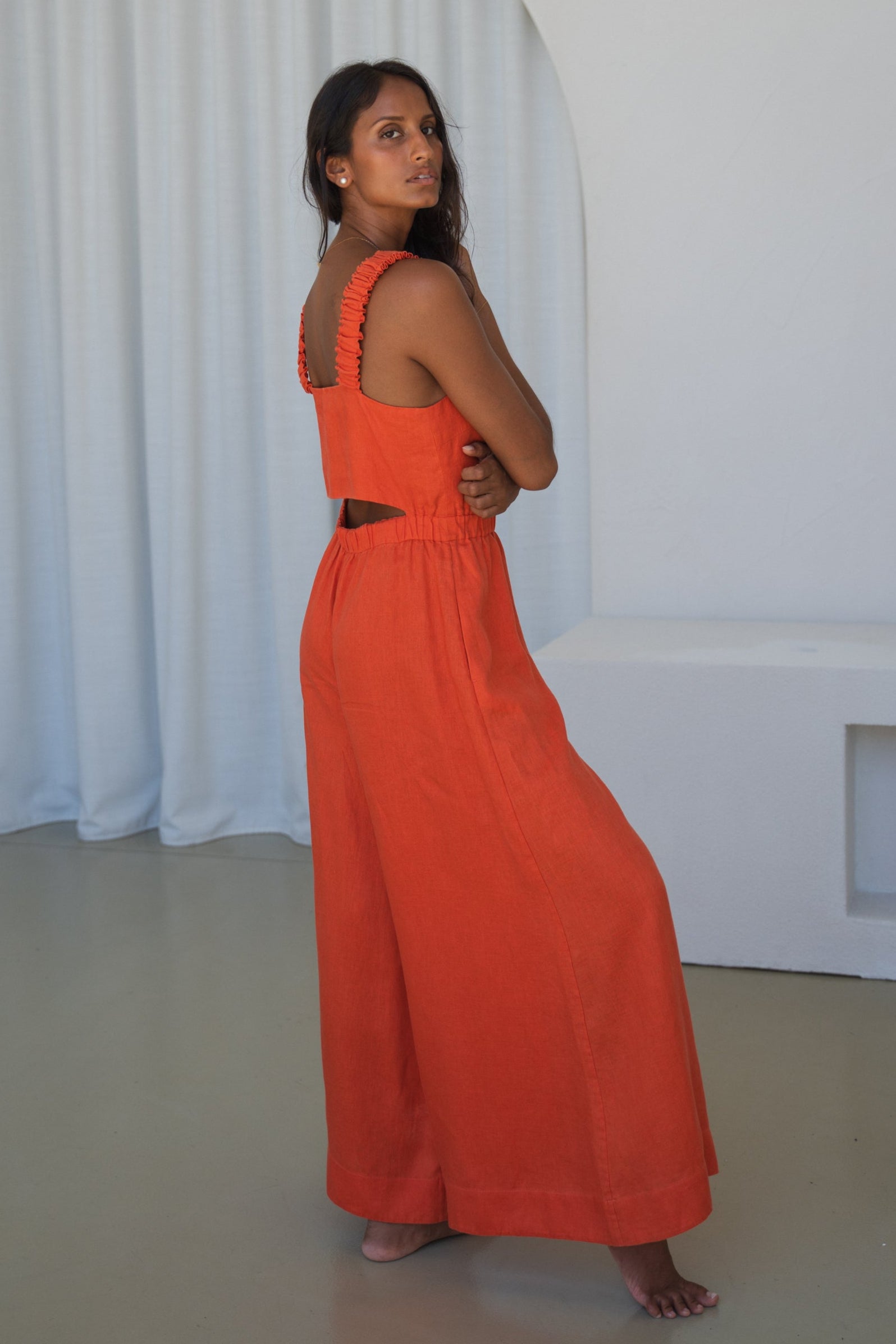 LILLY PILLY woman in an spritz Keely linen jumpsuit back view standing against a white curtain background
