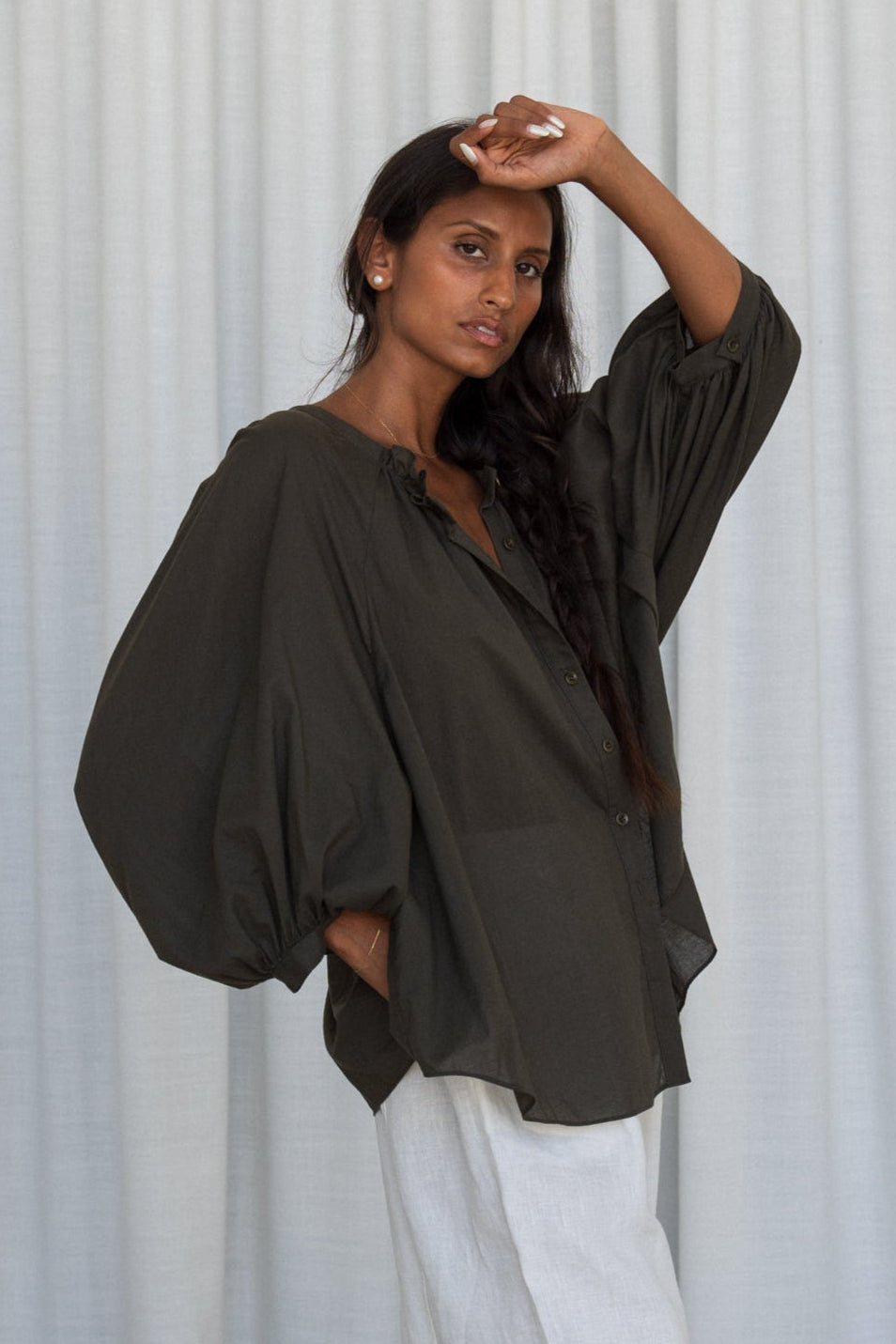 LILLY PILLY woman wearing a khaki Thalia cotton voile shirt blouse showing side view in front of a curtain background