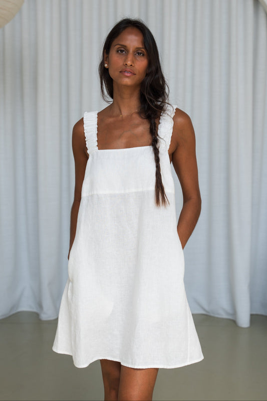 LILLY PILLY woman in an ivory Zahra linen dress showing back view while standing in front of a white curtain background
